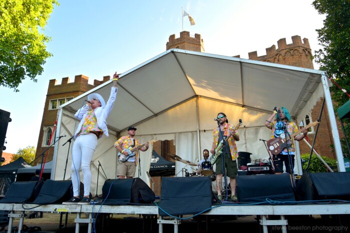 A man dressed all in white is on stage with his band, under a white canopy in front of hertford castle. It's a shot from the Rock at the Castle
