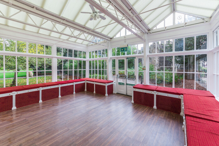 A side view of inside the conservatory with its white frame and red cushions on the benches around the sides.