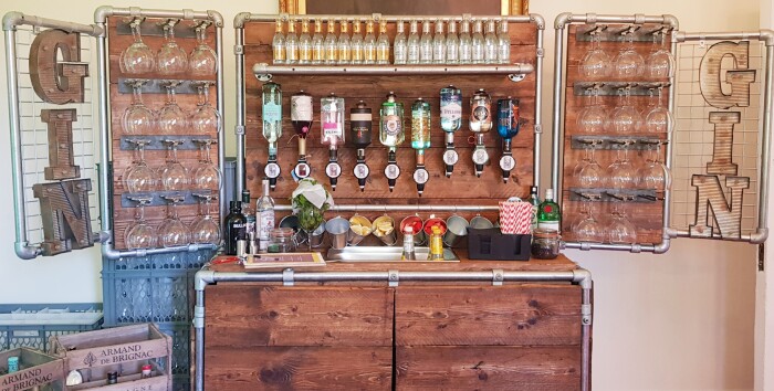 A wooden bar set up in a industrial style with glasses hanging on the side panel ans the words gin spelled out on the wings, bottles of of alcohol han