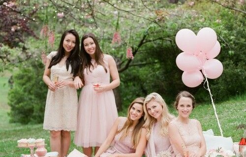 A hen party having a picnic, they are all dressed in pale pink sat and stood around a pink blanket with food and balloons