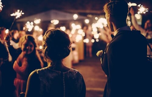A night time shot of a bride and groom in silhouette walking towards their friends and family in the background waving sparklers