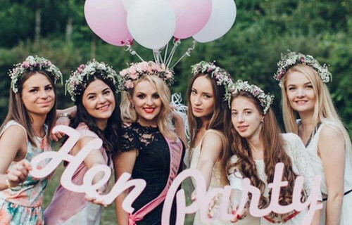 A hen group look at the camera holding a sign that says Hen party. with pink balloons in the background