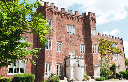 A side angle shot of Hertford Castle with blue skies above and green trees to the left