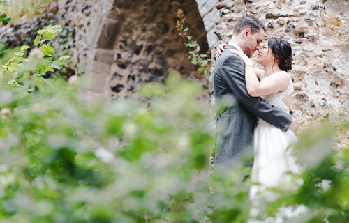 A couple hugging in front of the flint curtain wall with greenery in the foreground