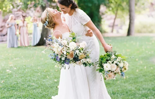 Two brides, hugging, clutching huge bouquets of blue, white and pink flowers on a grassy lawn