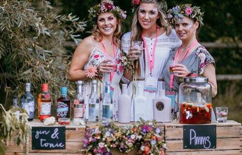 A bride with two bridesmaids stand behind a wooden bar holding drinks, lets of bottles are on the top of the bar, and the women all have flower crowns