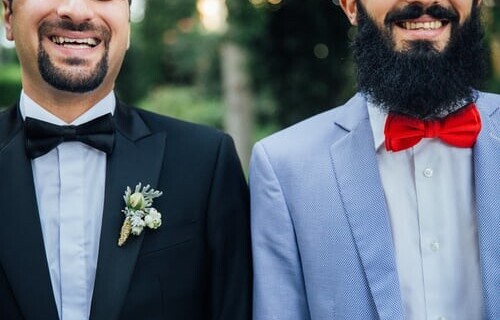Two breaded grooms, smile stood side by side, one has a red bow tie and blue jacket the other a black bow tie and jacket.