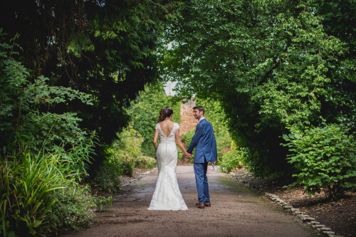 A bride and groom holding hands walking away from the camera under an archway of trees