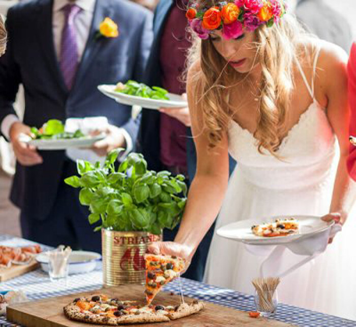 A bride leaning over a catering table grabbing a slice of pizza, she's wearing a flower crown of red flowers and a spaghetti strap dress