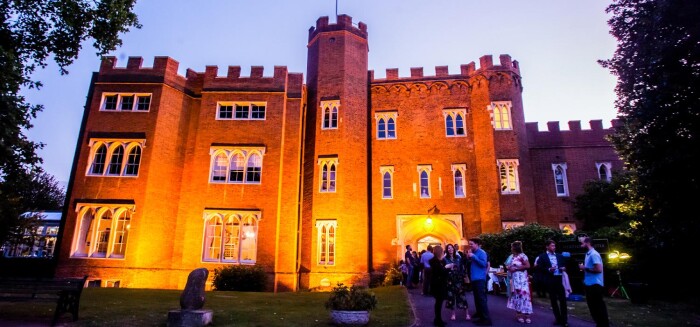 Hertford Castle at dusk, lit up by up lighters whilst guests enjoy drinks outside.