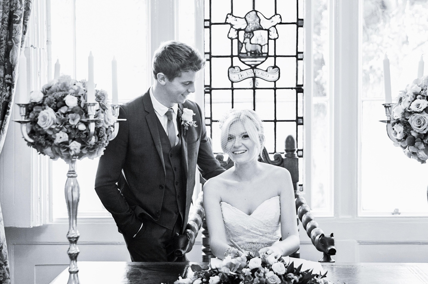 A man looks lovingly at his bride who is sat in the winged chair in front of the stained glass window