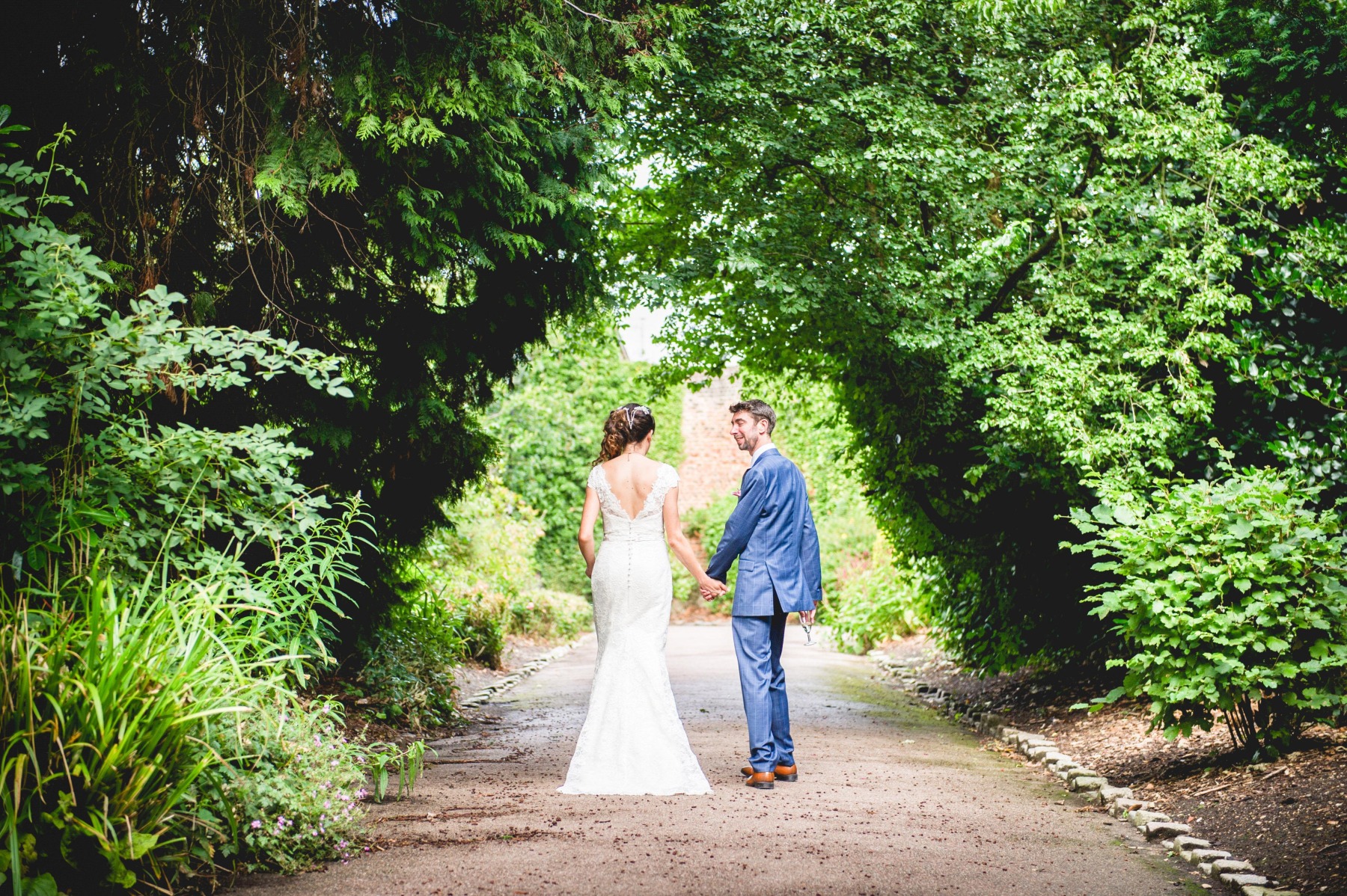 A bride and groom walking away from the camera on a path under an arch of green trees