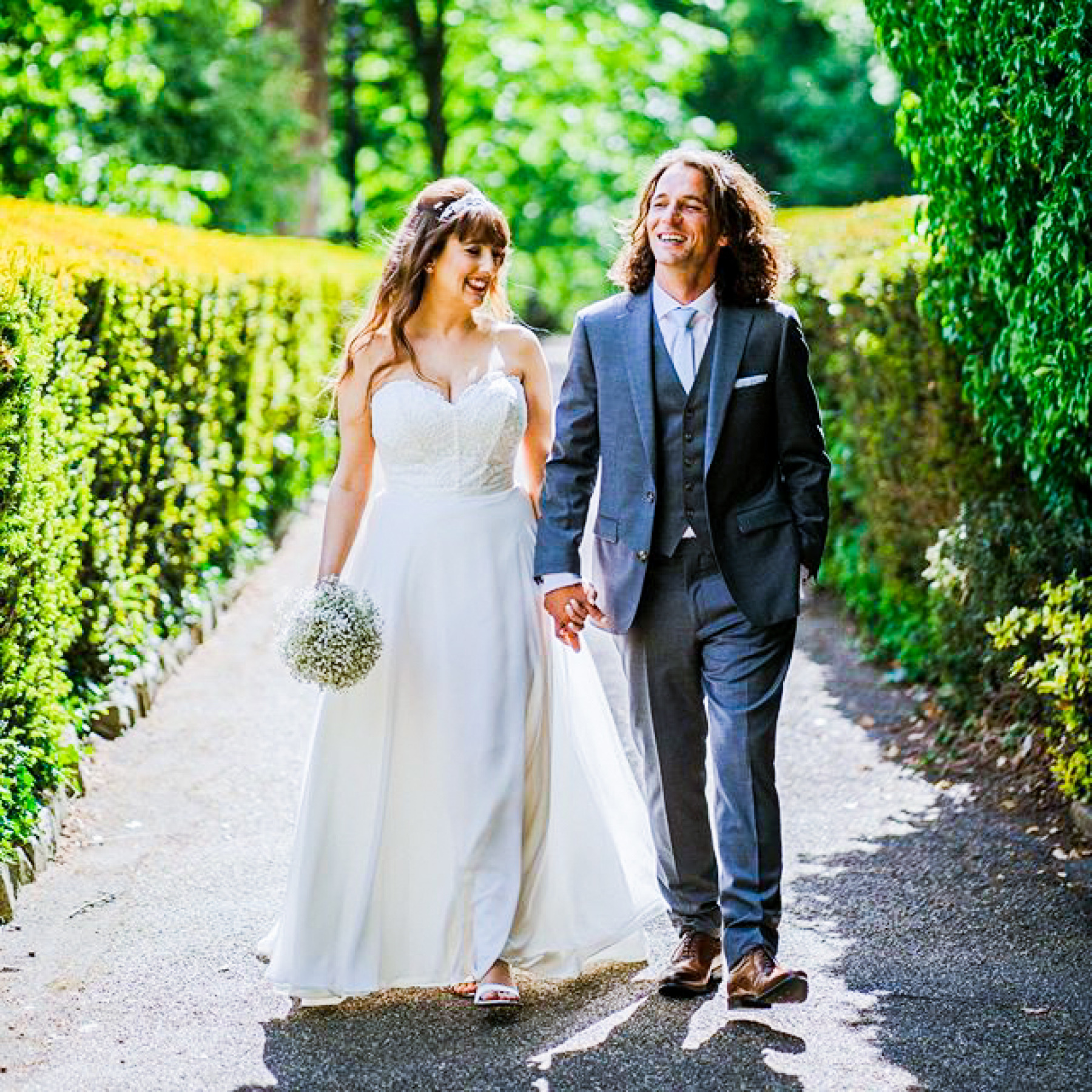 A bride and groom holding hands walking down the path towards the camera, flanked by hedges