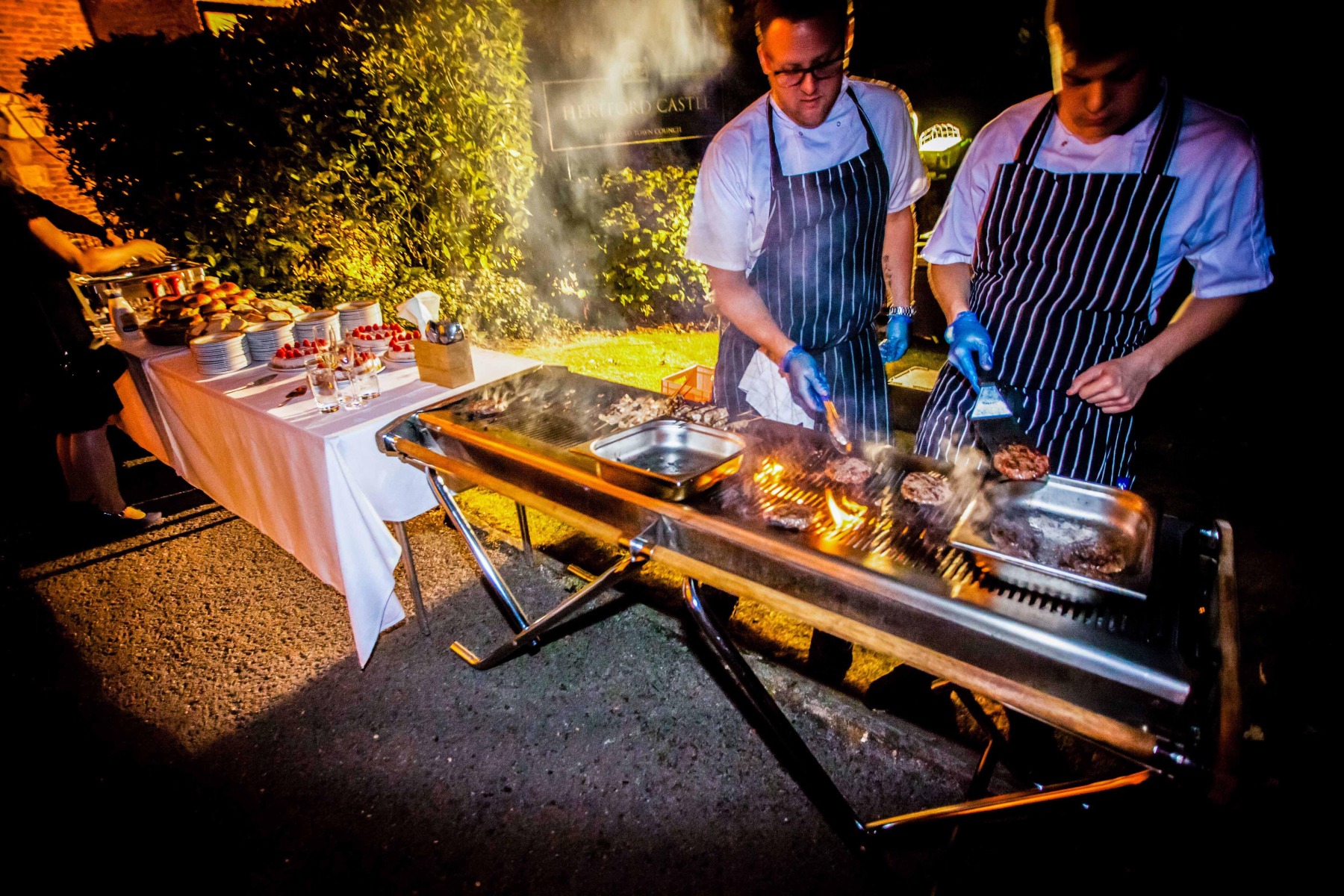 A close up of caterers cooking over an open flamed BBQ at night