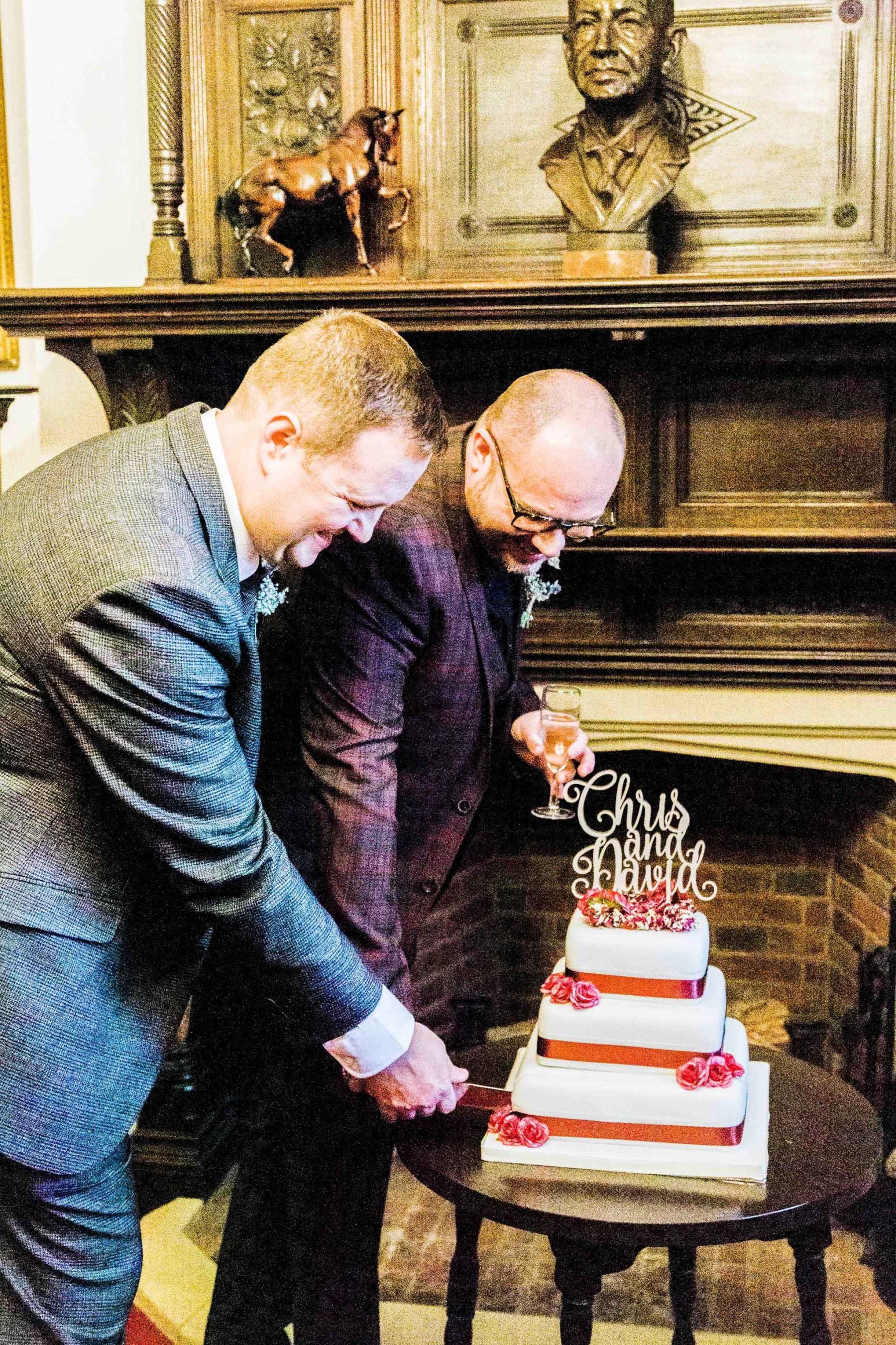 A couple wearing a maroon suit and a grey suit cutting their wedding cake together