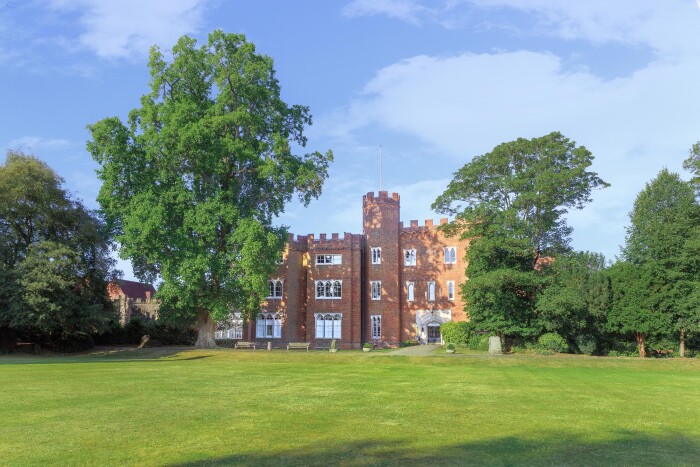 Hertford Castle viewed from the main lawn, surrounded by trees, with a blue sky above