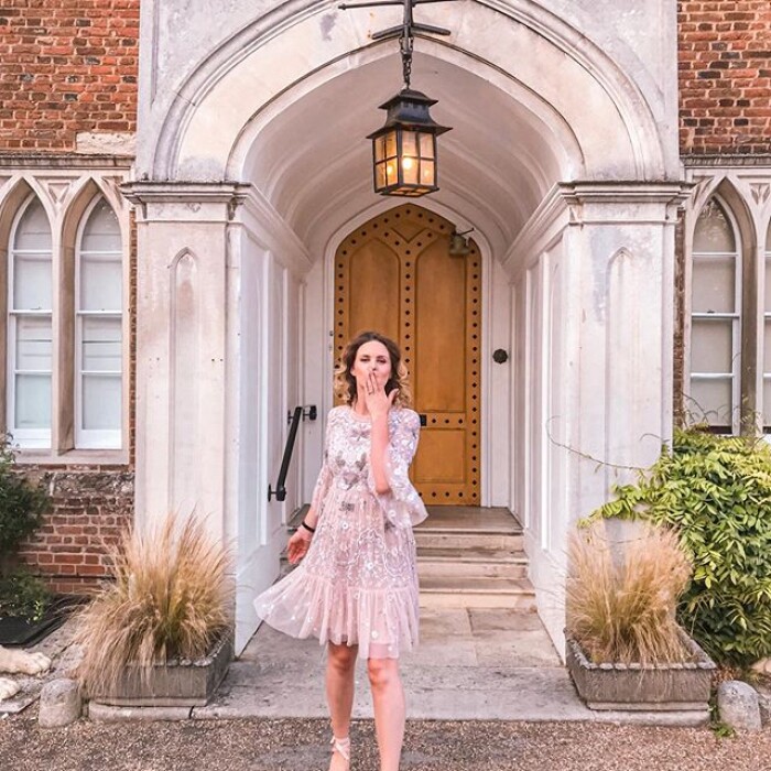 A woman is stood outside the wooden door and white stone arch of Hertford castle in a pink floaty dress, blowing a kiss to the camera