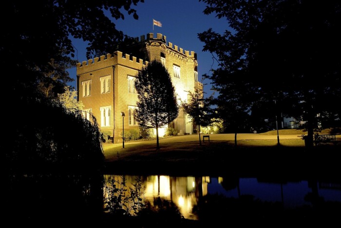 A side view of Hertford Castle at night, light up by the uplighters, the trees silhouettes in the foreground