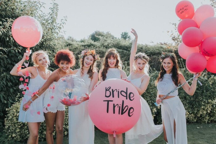 A hen party, all dressed in white posing in front of the camera with pink balloons. One large one in the middle says Bride Tribe on it