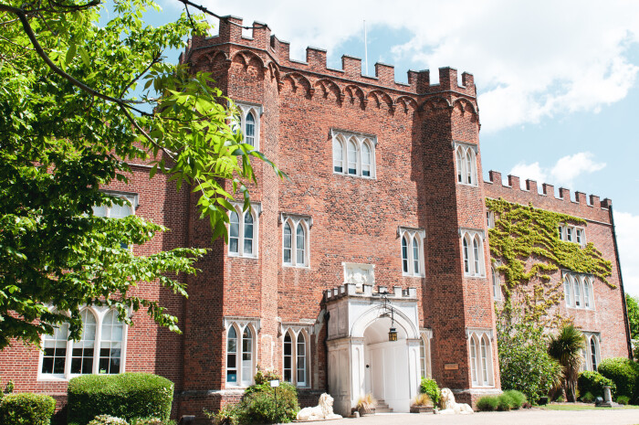 A side angle view of the front of Hertford Castle with blue skies above and a green tree in the foreground