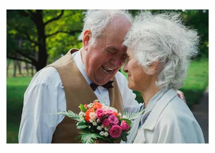 An older couple, the lady holding a bunch of pink and orange flowers, their heads meet lovingly as they laugh