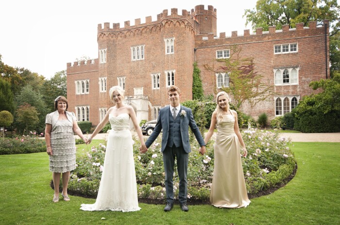 A wide shot of Hertford Castle with a bride, groom, bridesmaid and mother of the bride in the rose gardens holding hands