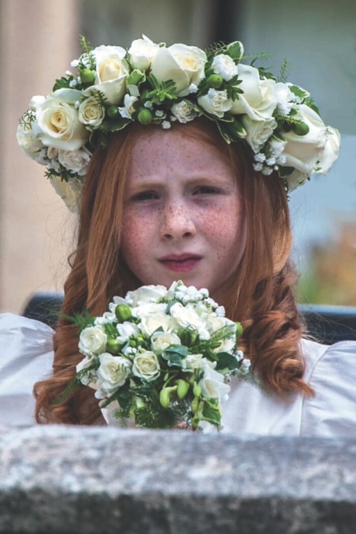A red headed flower girl, holding a bouquet of white flowers, whilst wearing a flower crown