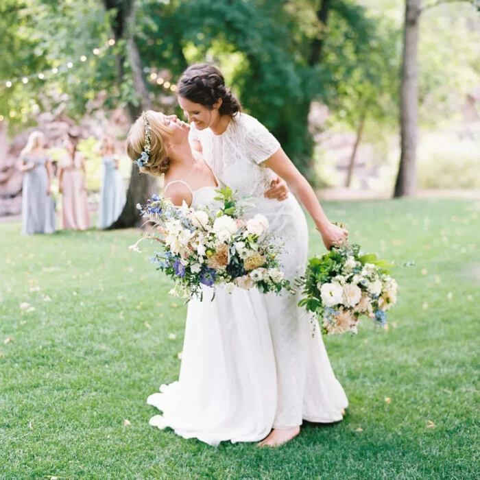 Two brides, hugging, clutching huge bouquets of blue, white and pink flowers on a grassy lawn