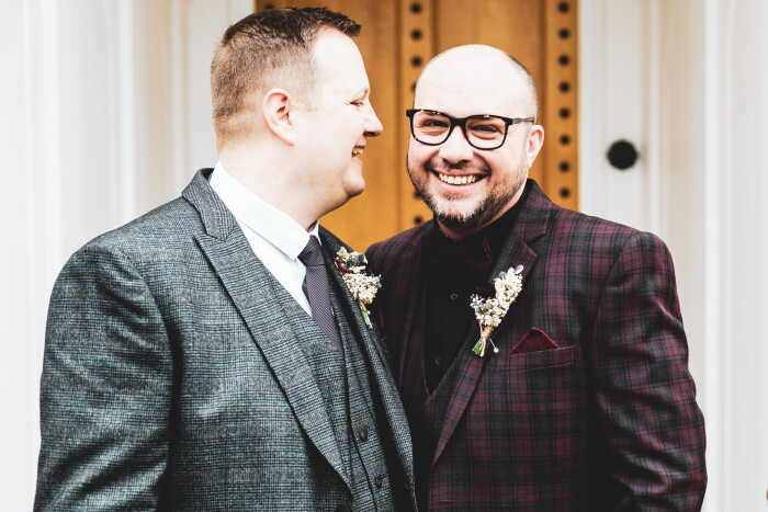 Two grooms stand outside the castle's wooden door, one looking at the camera smiling, the other looking at his husband also smiling