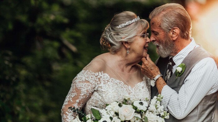 An older couple, in wedding attire, eskimo kissing, the groom gently touching the brides chin.