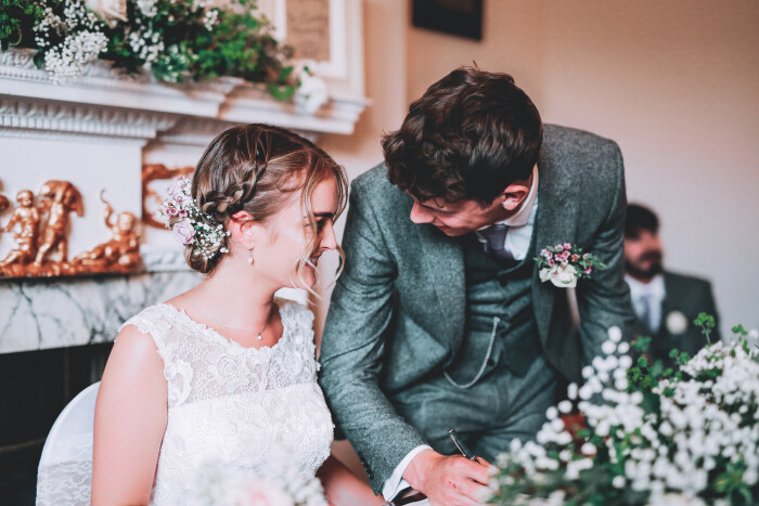 A bride and groom are signing the register in front of the ornate fireplace in the Salisbury room. The groom is looking lovingly at his bride.