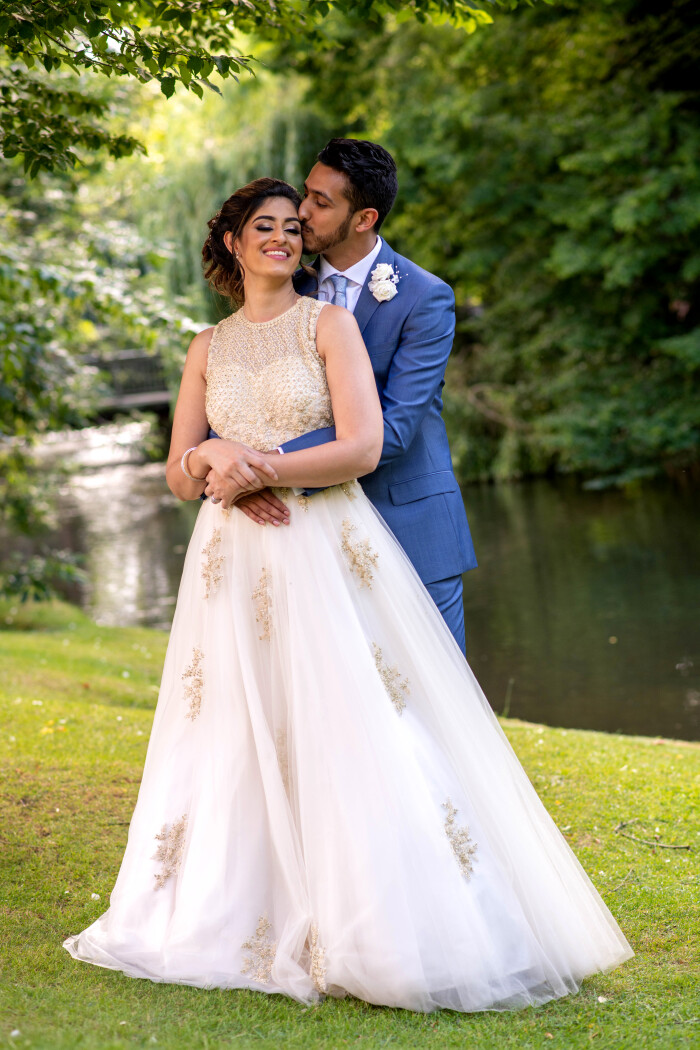 A groom in a bright blue suit, holding his bride round the waist, kissing the side of her head as she smiles at the camera, the river lea in the backg