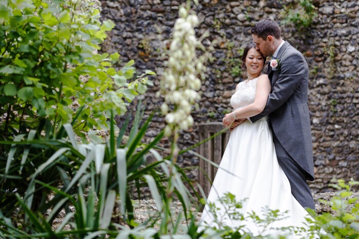 A couple hugging in front of the flint curtain wall, the groom kissing the bride on the head, with greenery in the foreground